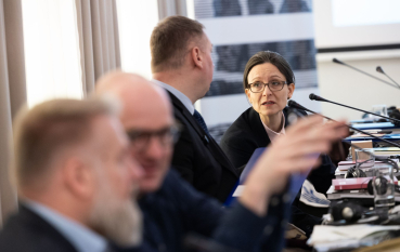 Grupa osób przy stole z mikrofonami, zbliżenie na kobietę w okularach. 