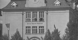 Czarno-biała fotografia przedstawiająca front budynku koszarowego z napisem „Julius Schreck-Kaserne”. Przed wejściem widoczne są flagi z symboliką nazistowską, wartownik oraz przechodnie na ulicy
