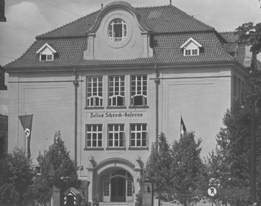 Czarno-biała fotografia przedstawiająca front budynku koszarowego z napisem „Julius Schreck-Kaserne”. Przed wejściem widoczne są flagi z symboliką nazistowską, wartownik oraz przechodnie na ulicy