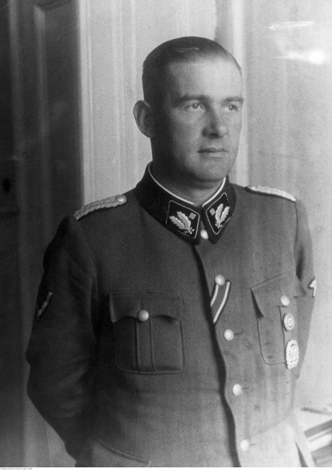 Czarno-biały portret mężczyzny w mundurze SS stojącego bokiem przy oknie. Postać ma krótko ostrzyżone włosy i patrzy w dal; na kołnierzu widoczne są oznaczenia stopnia oraz elementy umundurowania charakterystyczne dla okresu II wojny światowej.