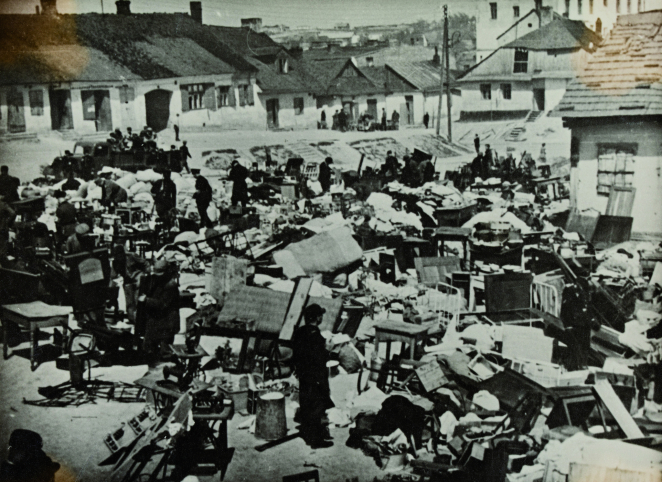 Zdjęcie czarno-białe. Porozrzucane meble i inne przedmioty na placu. Dookoła niskie zabudowania. Między rzeczami przechadzają się ubrani na czarno mężczyźni