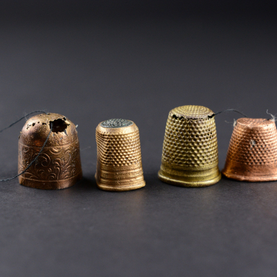 A row of four sewing thimbles placed side by side against a dark background. They are made of metals in different shades: copper, brass, and gold. One of them, on the left, is richly decorated with a floral motif and has a visible crack and a missing fragment at the top. The others have a classic dotted texture.