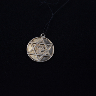 A round, silver-colored medallion photographed centrally against a black background. Inside the circle is a Star of David, with a Hebrew inscription at its center.