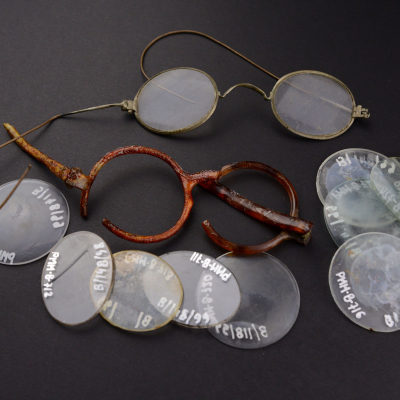On a dark background lie two pairs of damaged eyeglass frames – one metal and wire, the other made of dark amber-colored plastic. Next to them are scattered several round glass lenses.