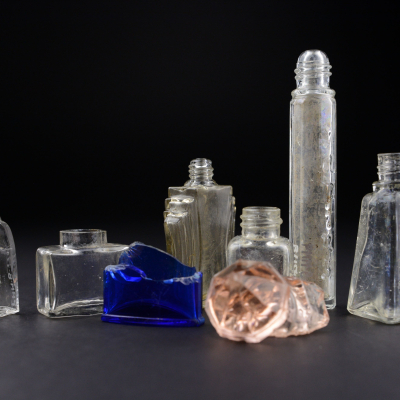 Six glass bottles of various shapes – from slender and tall to short and bulbous – arranged on a black background. One is made of deep blue glass, another has a pink hue. Some of the vessels are chipped and incomplete.