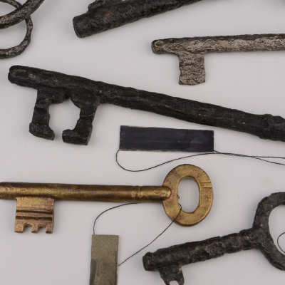 Several old metal keys of various sizes and shapes laid out on a light background. Some keys are covered with a thick layer of dark rust and have decorative handles, while others are smaller and made of brass. Black strings with museum labels are attached to the keys