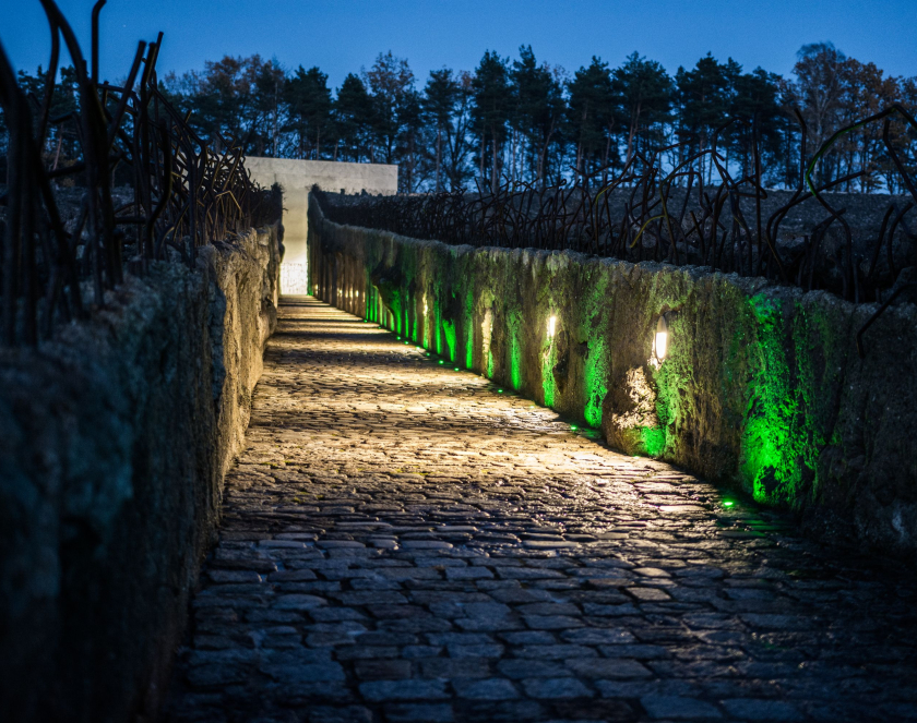 Night photograph. A cobblestone path flanked by illuminated walls and iron bars. Trees are visible in the background.