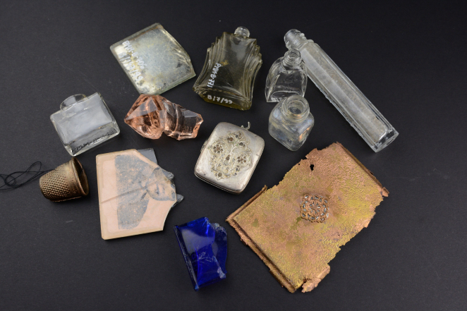 Top-down view of a group of various objects on a dark background. Among them are glass bottles from perfumes or medicines, a silver engraved powder box, a fragment of a child’s photograph on glass, blue glass, and a metal piece shaped like a notebook cover.