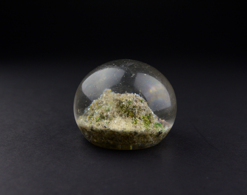 Small, transparent glass object shaped like a flattened sphere, resembling a paperweight or decorative element. Inside the glass is an irregular, textured mass in earthy, green, and white colors, resembling a miniature landscape or solidified sand. The object is photographed centrally against a dark, matte background.
