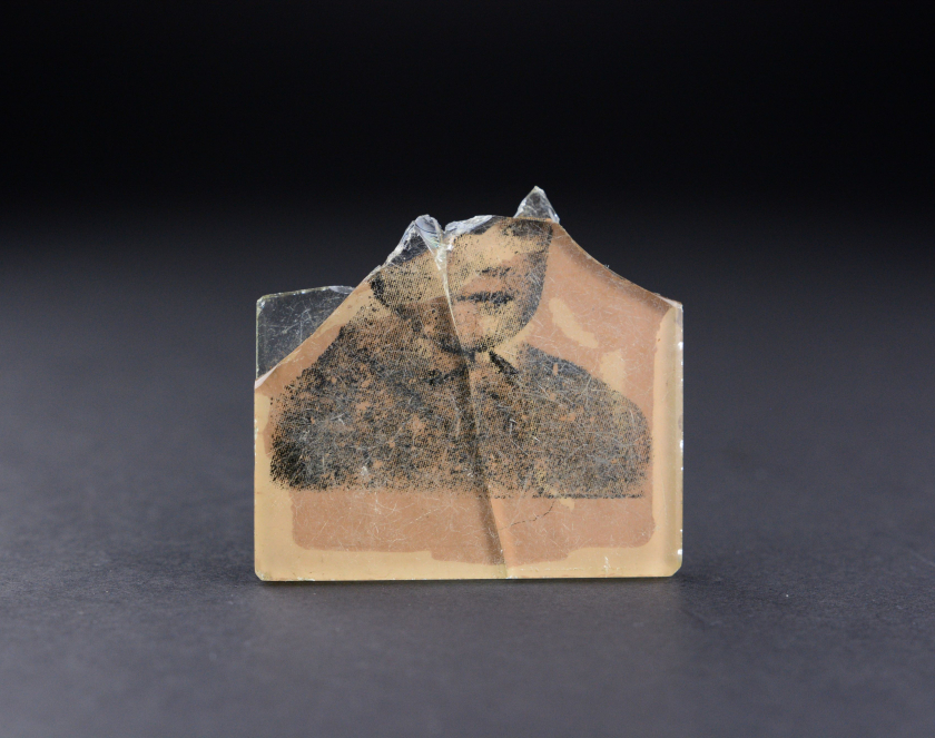 Small, chipped piece of a mirror with a black-and-white photograph showing the lower part of a face and a child’s bust. The artifact rests on a dark background.