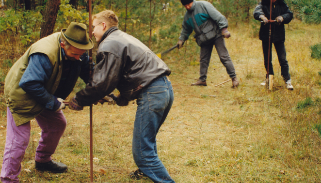 Archival color photograph from 1997 showing archaeologists and staff conducting test boreholes in the forest on the site of the former Bełżec camp. The men are using hand-operated geological augers to examine soil layers.