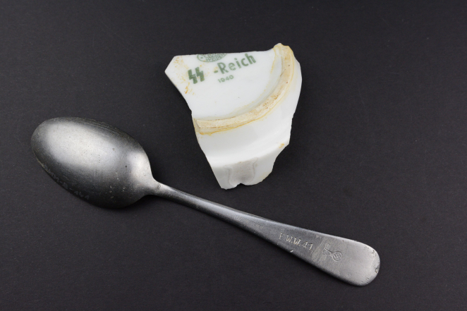 On a dark background lie two objects: a matte metal spoon and a white ceramic fragment of a plate bottom. The handle of the spoon bears an engraving of an eagle holding a swastika and the mark ‘F.W.W. 41’. The ceramic fragment shows a green print with two lightning bolts (SS runes), the inscription ‘-Reich,’ and the date ‘1940’.