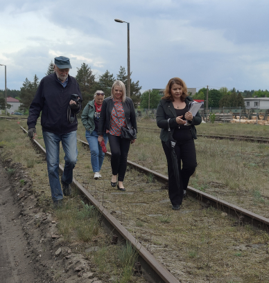 Grupa czterech dorosłych osób idzie wzdłuż nieczynnego, porośniętego trawą toru kolejowego pod zachmurzonym niebem. Po prawej stronie w tle widoczne są ułożone bele drewna oraz budynki.