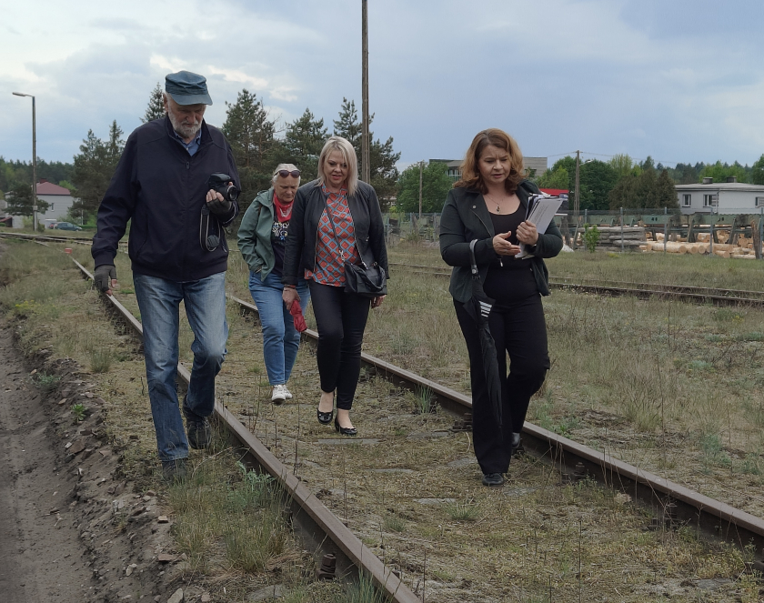 Grupa czterech dorosłych osób idzie wzdłuż nieczynnego, porośniętego trawą toru kolejowego pod zachmurzonym niebem. Po prawej stronie w tle widoczne są ułożone bele drewna oraz budynki.