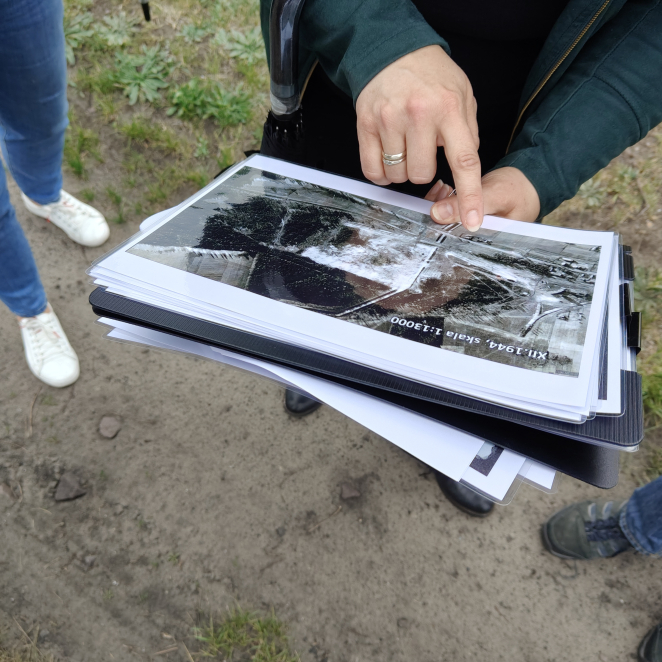 Bliskie ujęcie dłoni kobiety wskazującej palcem na archiwalne zdjęcie lotnicze z podpisem „XII.1944, skala 1:13000”. Kobieta trzyma w rękach czarną teczkę z dokumentami, w tle widoczne są nogi innych osób stojących na ziemi.