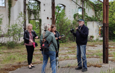 Cztery osoby stoją na betonowym podłożu wewnątrz zrujnowanego budynku bez dachu, z widoczną metalową konstrukcją sufitu. Starszy mężczyzna po prawej stronie gestykuluje i rozmawia z pozostałymi uczestnikami. Ściany z cegły i tynku są porośnięte bluszczem i pokryte graffiti.