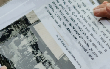 Zbliżenie na trzymaną w dłoniach kartkę papieru w plastikowej koszulce. Na stronie znajduje się tekst w języku polskim dotyczący śmiertelności w obozie i relacji świadka, a po lewej stronie umieszczono małe, czarno-białe archiwalne zdjęcie z podpisem zawierającym nazwę „Bełżec 1940”.