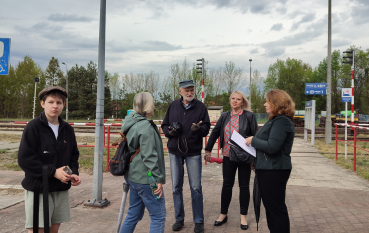 Grupa pięciu osób rozmawia na brukowanym peronie kolejowym pod chmurnym niebem. W tle widoczne są tory kolejowe, czerwone barierki ochronne, semafory oraz niebieska tablica informacyjna stacji.