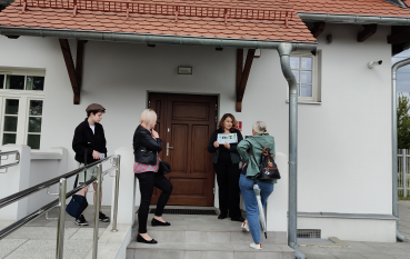 Grupa osób stoi przed wejściem do odnowionego, białego budynku z dachem z czerwonej dachówki. Jedna z kobiet stoi na podeście przed brązowymi drzwiami i pokazuje pozostałym trzymaną w rękach kartkę. Po lewej stronie widoczna jest rampa z metalową barierką.