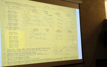 Ekran prezentacji przedstawiający fragment historycznego kwestionariusza wypełnionego odręcznym pismem. Widoczne są pola dotyczące narodowości i pochodzenia.