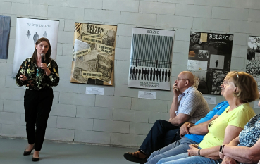 Wnętrze sali z ceglanymi ścianami, na których wiszą liczne plakaty i obrazy związane z tematyką historyczną obozu w Bełżcu. Widoczni są słuchacze siedzący w rzędach krzeseł. Po lewej stornie prelegentka z mikrofonem.