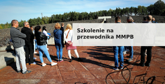 Promotional banner for the guide training course at the Museum and Memorial Site in Bełżec. The photo shows a group of people standing on the grounds of the former death camp, with a stony field, a memorial wall, and a forest visible in the background. The image displays the text "Szkolenie na przewodnika MMPB" ("Guide Training – MMPB") in bold black lettering on a light, semi-transparent background.