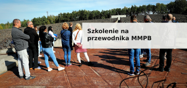 Baner promocyjny szkolenia dla przewodników Muzeum i Miejsca Pamięci w Bełżcu. Na zdjęciu grupa osób stojąca na terenie dawnego obozu zagłady, w tle kamienisty teren i ściana pamięci oraz las. Na zdjęciu widoczny jest napis „Szkolenie na przewodnika MMPB” w czarnej, pogrubionej czcionce na jasnym, półprzezroczystym tle. 