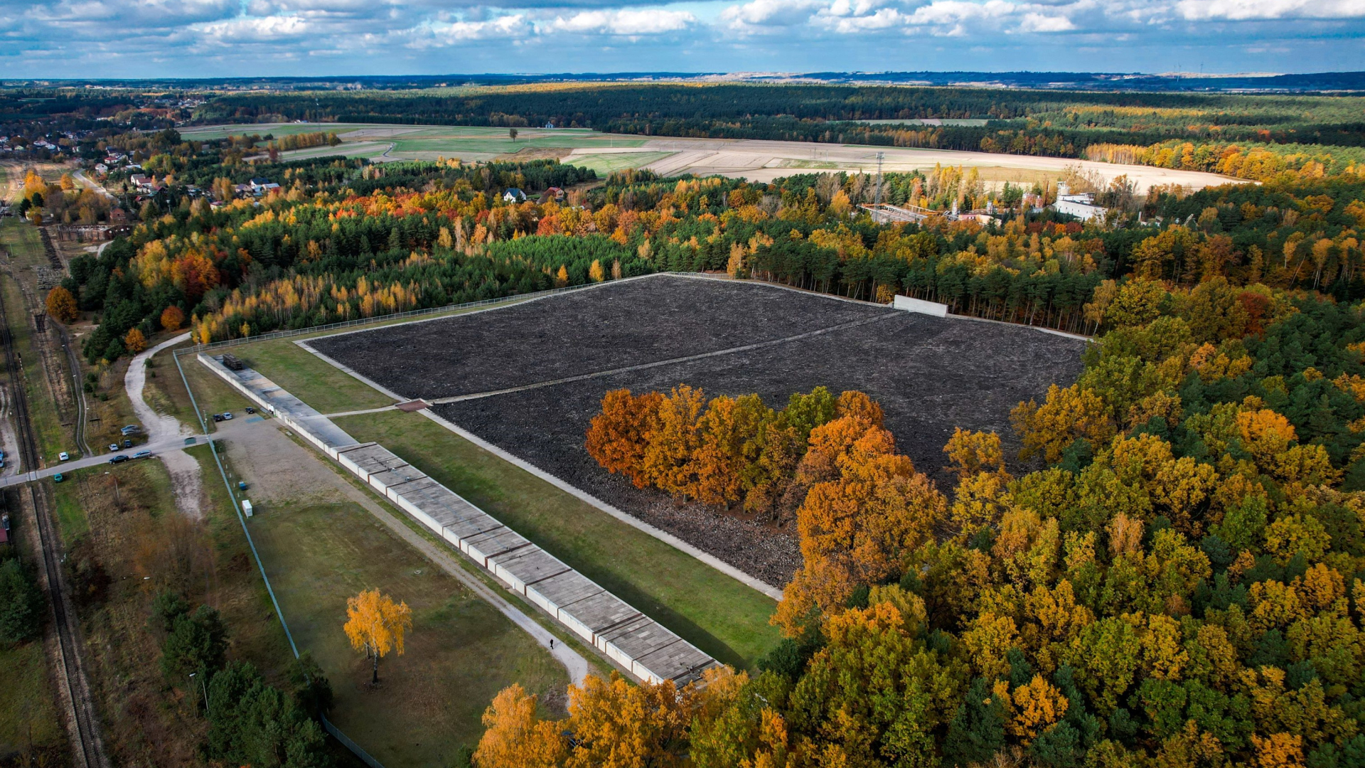 Zdjęcie lotnicze przedstawiające teren Muzeum i Miejsca Pamięci w Bełżcu jesienią. W centralnej części widoczne duże, prostokątne pole pokryte ciemnym żużlem i kamieniami, otoczone niskim betonowym chodnikiem. Wzdłuż jednej z krawędzi ciągnie się długi, betonowy budynek muzeum. Teren otoczony jest lasem mieniącym się jesiennymi barwami – zielenią, żółcią i pomarańczem. W tle widoczne pola uprawne, zabudowania wsi oraz linia horyzontu pod błękitnym niebem z białymi chmurami.