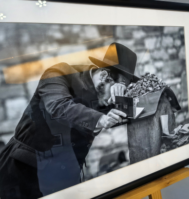 Oprawiona fotografia czarno-biała: mężczyzna w kapeluszu, pochylony nad nagrobkiem, trzyma w dłoni mały modlitewnik; na płycie nagrobnej leży sterta kamyków, w tle rozmyty mur cmentarny. W szklanej taflie odbijają się fragmenty sali wystawowej.