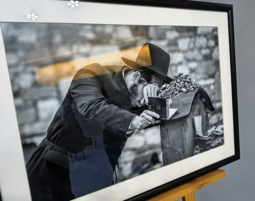 Oprawiona fotografia czarno-biała: mężczyzna w kapeluszu, pochylony nad nagrobkiem, trzyma w dłoni mały modlitewnik; na płycie nagrobnej leży sterta kamyków, w tle rozmyty mur cmentarny. W szklanej taflie odbijają się fragmenty sali wystawowej.