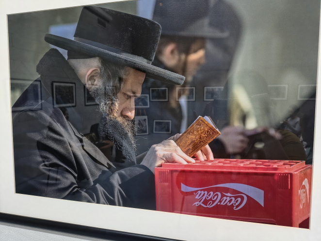 Oprawiona w ramę fotografia z wystawy: mężczyzna w czarnym kapeluszu i długim płaszczu, z siwiejącą brodą i pejsami, trzyma w dłoni oprawiony w skórę modlitewnik ozdobiony tłoczonym wzorem, opierając go o czerwoną skrzynkę po Coca-Coli. W tle, rozmyta, widoczna druga postać w podobnym stroju. W szybie odbijają się inne fotografie z galerii.