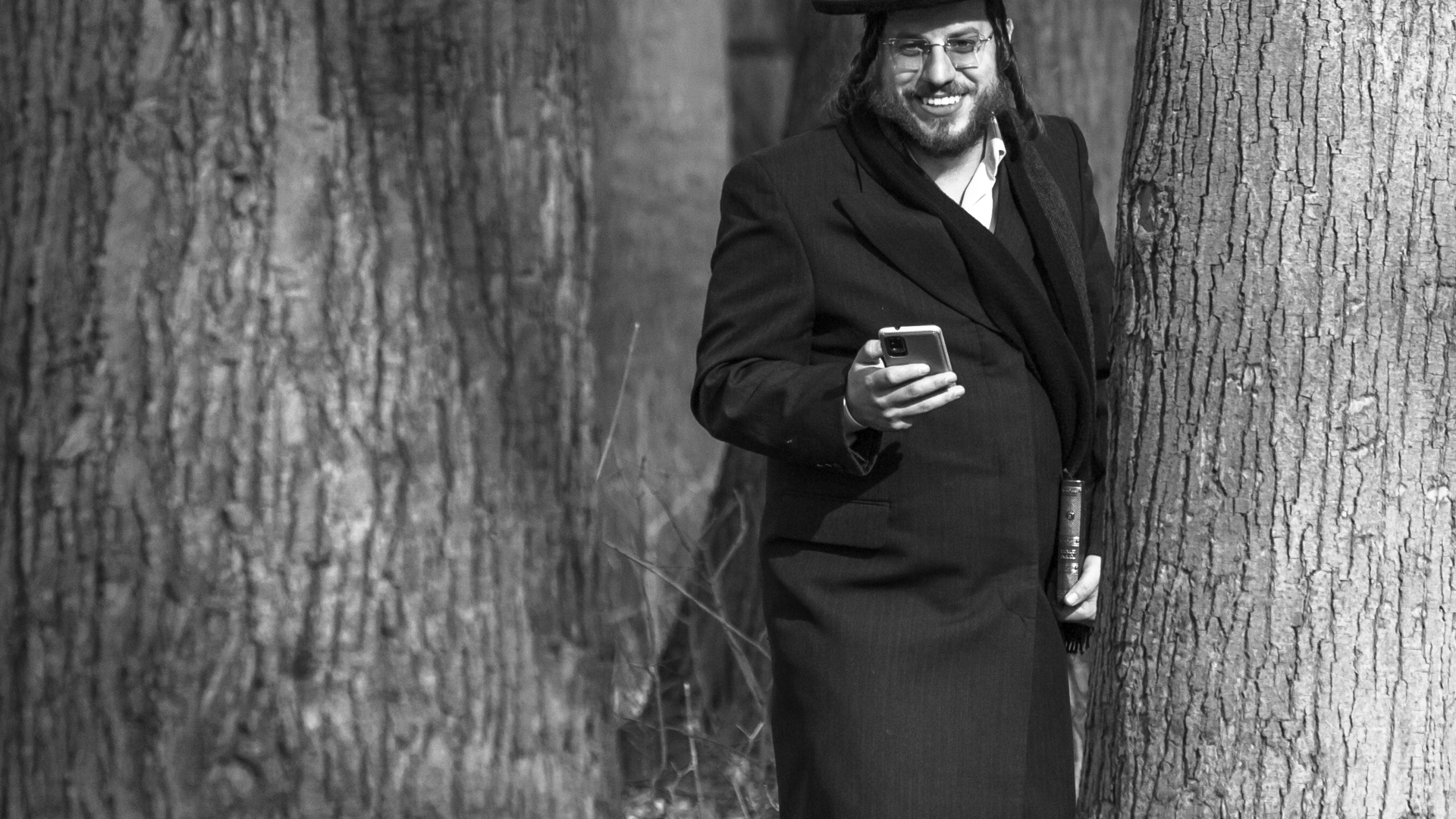 Czarno-biała fotografia: młody mężczyzna w chasydzkim stroju i kapeluszu, z pejsami, stoi uśmiechnięty między dwoma grubymi pniami drzew, trzymając w jednej dłoni telefon, a w drugiej modlitewnik.