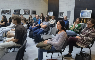 Publiczność siedząca w rzędach podczas spotkania, w tle ściana z oprawionymi fotografiami.