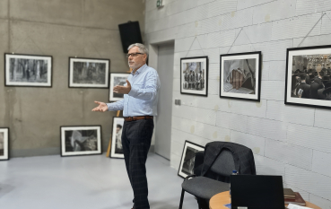 Siwowłosy mężczyzna w jasnoniebieskiej koszuli stoi samotnie w sali, gestykulując podczas opowieści; w tle liczne oprawione fotografie czarno-białe, obok krzesło i stolik z laptopem.