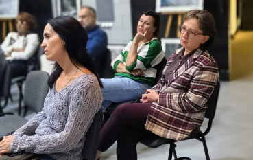 Trzy kobiety siedzące obok siebie, uważnie słuchające; w tle rozmyte fotografie z wystawy oraz złocisty akcent oświetlenia.
