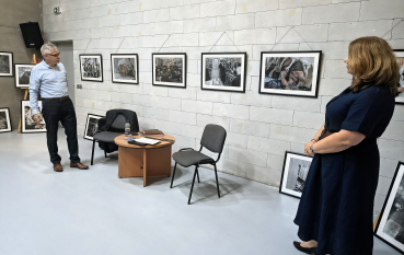Mężczyzna w jasnej koszuli stoi po lewej stronie sali, a kobieta w granatowej sukience ogląda rząd pięciu oprawionych fotografii zawieszonych na ceglanej ścianie; obok stolik z krzesłami i dodatkowe prace oparte o ścianę.