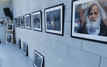 Widok ściany z rzędem oprawionych fotografii portretowych zawieszonych na jasnej ceglanej ścianie; na pierwszym planie duży portret starszego mężczyzny z siwą brodą, część prac oparta dodatkowo o podłogę.