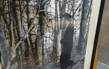 Oprawiona fotografia ujęta pod kątem: sylwetka mężczyzny w długim czarnym płaszczu i kapeluszu, stojącego samotnie wśród wysokich, bezlistnych drzew, w stonowanym, zimowym świetle.
