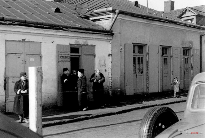 Czarno-biała fotografia przedstawiająca fragment ulicy w Zamościu. Przed parterowymi budynkami stoją i rozmawiają mieszkańcy, wśród nich mężczyźni w tradycyjnych żydowskich nakryciach głowy. Po prawej stronie widoczny fragment samochodu.