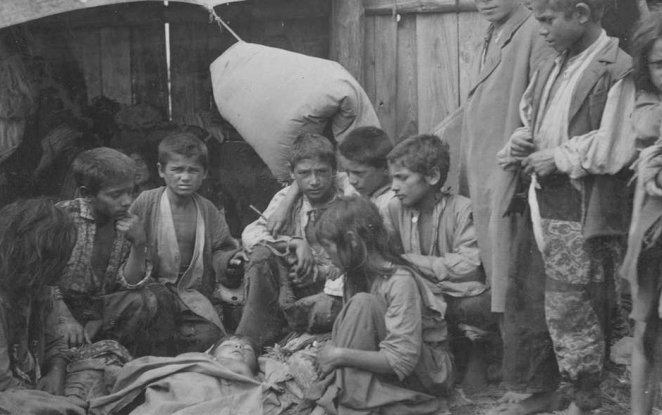 Czarno-biała fotografia archiwalna przedstawiająca grupę romskich i sintyjskich dzieci w obozie pracy w Bełżcu. Dzieci siedzą i stoją wokół osoby leżącej na ziemi. W tle widoczne są namioty i drewniane ogrodzenie.